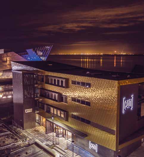 Aerial night view of a modern building with a gold facade and illuminated signs by a body of water, with city lights in the distance.