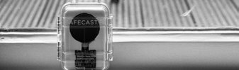 Close-up black and white photo of a clear plastic box labeled "AFECAST" containing a circular black electronic component.