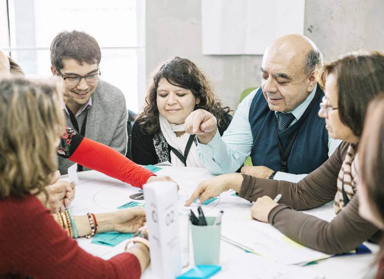 A group of diverse people gathered around a table, actively pointing at and discussing a diagram or document laid out in the center.