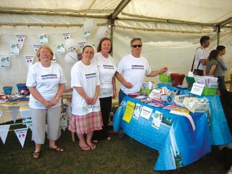 Four people in matching t-shirts standing behind a stall under a tent, displaying items at a community event.