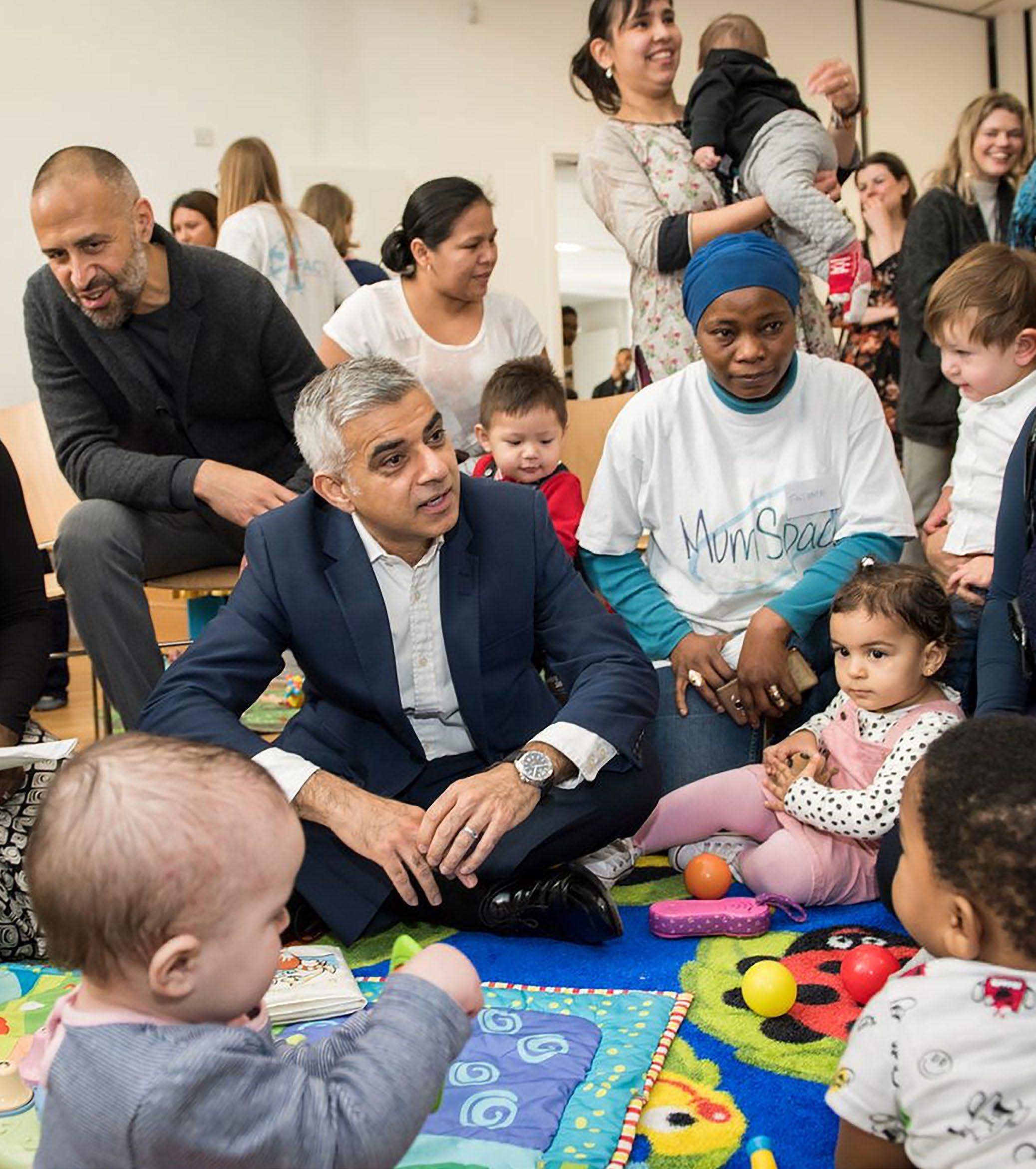 A group of adults and young children sitting on a colorful mat, including a public figure interacting with them.