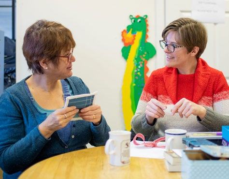 Two women smiling and talking at a table, one holding a small item. A colorful dragon decoration is in the background.