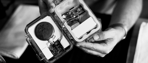 Hands holding an open, clear case revealing internal electronic components, including a large circular sensor.