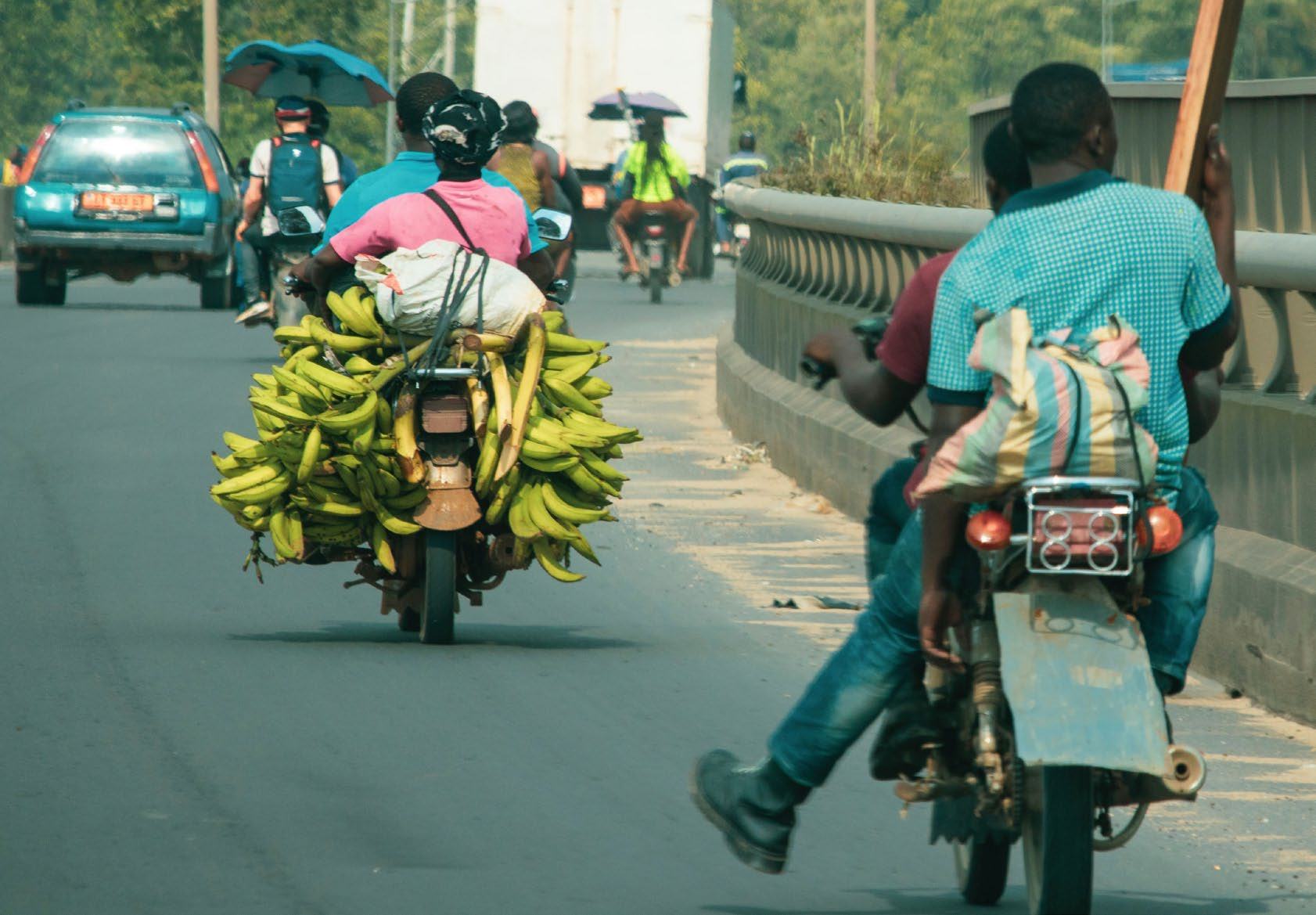 A street scene with two motorcycles, one heavily loaded with green bananas, and another carrying two people, depicting local transportation.