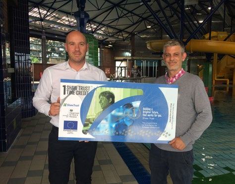 Two men standing by a swimming pool holding a large display board from "Shaw Trust 1 Time Credit" and the European Social Fund.