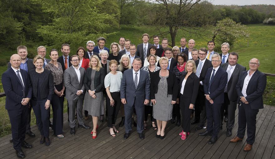 A large group photo of diverse business professionals standing on a wooden deck outdoors with trees and green landscape in the background.