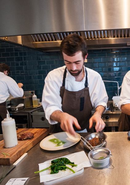 Chef in a white shirt and brown apron plating a dish with spinach in a busy professional kitchen.