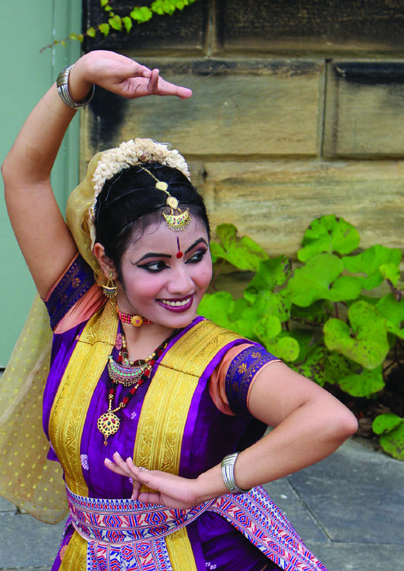 A woman in traditional Indian dress, including a purple and gold outfit and jewelry, performs a dance pose outdoors.