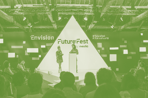 Two speakers present on a stage at the "Envision FutureFest" event, with a large triangular light projection and an audience present.