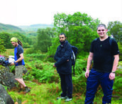 Three men in outdoor clothing stand in a bushy, hilly landscape, possibly on a hike.