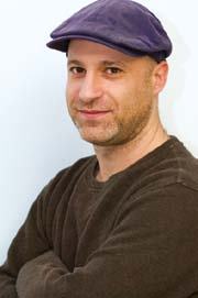Headshot of a smiling man with a beard, wearing a purple flat cap and a brown shirt.