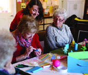 Three women, two elderly and one younger, are engaged in a crafting activity with colorful materials at a table.