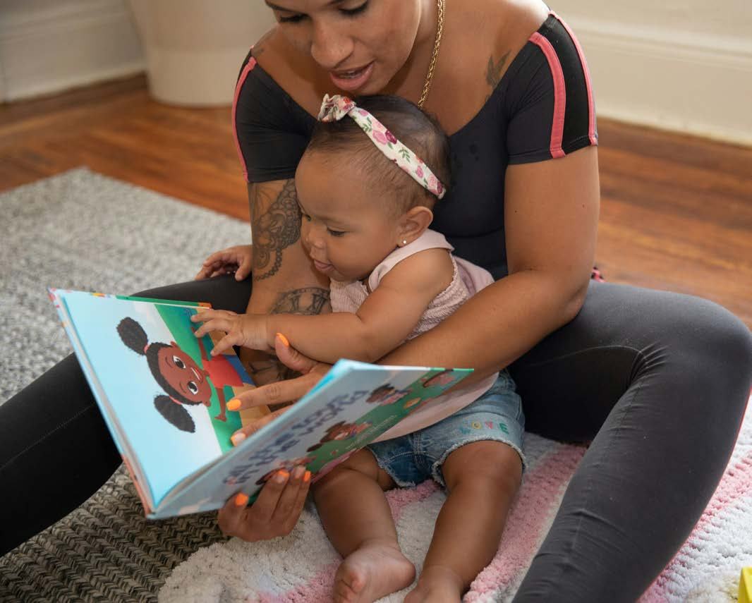 A woman with tattoos and a baby sit on the floor, looking at a colorful children's book with a smiling girl character.