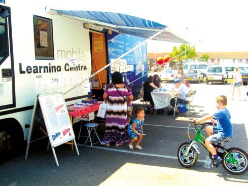 Mobile Learning Centre van with people around tables, and a child on a bike, at an outdoor community event.