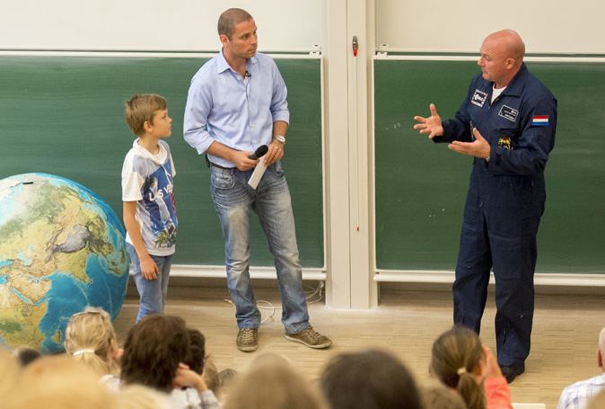 An astronaut in a flight suit speaks to a group of children and adults, with a large globe visible on the left.