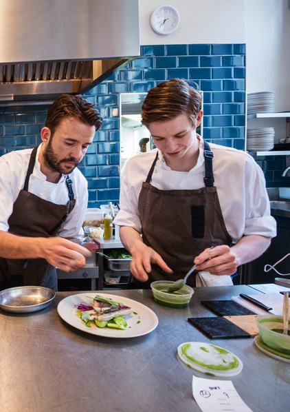 Two chefs in aprons meticulously plating dishes in a professional kitchen with blue tiled walls.