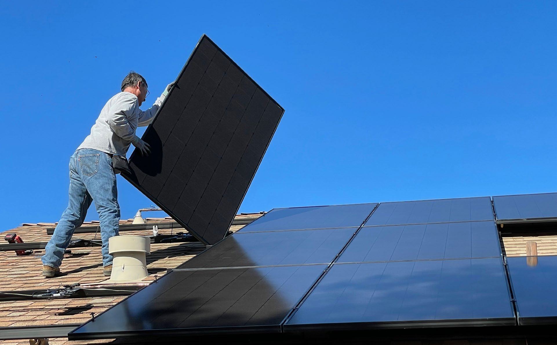 Worker on a roof installing a black solar panel onto a residential roof, with other panels already in place under a clear blue sky.