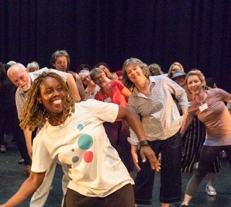 A group of adults smiling and leaning over in a dance or movement class, with a woman in the foreground wearing a logo t-shirt.