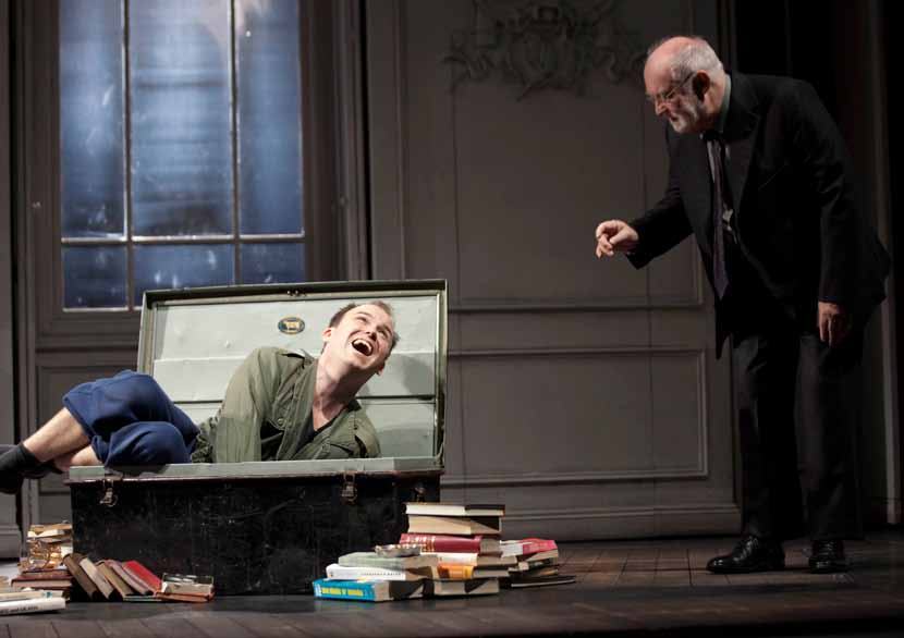 Two male actors on stage, one in a suit points at a younger man laughing inside an open trunk, surrounded by books.