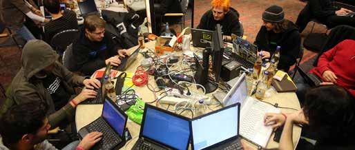Group of people collaborating around a cluttered table with multiple laptops and cables, possibly a hackathon.