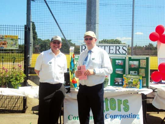 Two men from a community composting initiative stand behind a table with informational displays and red balloons at an outdoor event.