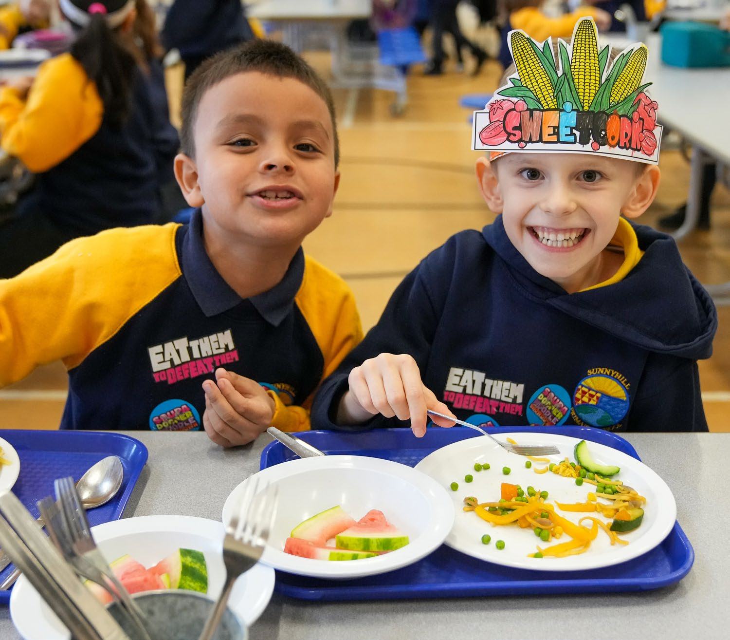 Two smiling primary school boys in 'Eat Them to Defeat Them' shirts eat vegetables and fruit at lunch. One boy wears a 'Sweet Corn' hat, illustrating a healthy eating campaign.