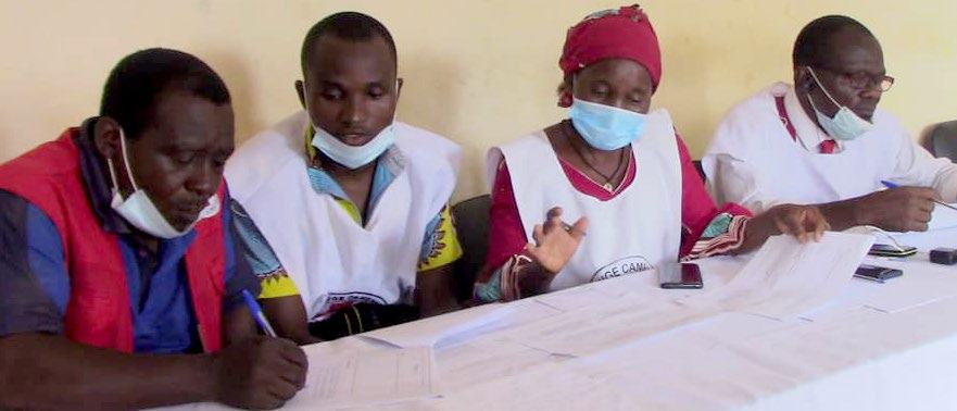 Four Red Cross volunteers, three wearing masks, seated at a table, reviewing documents and writing, likely in a meeting or work session.