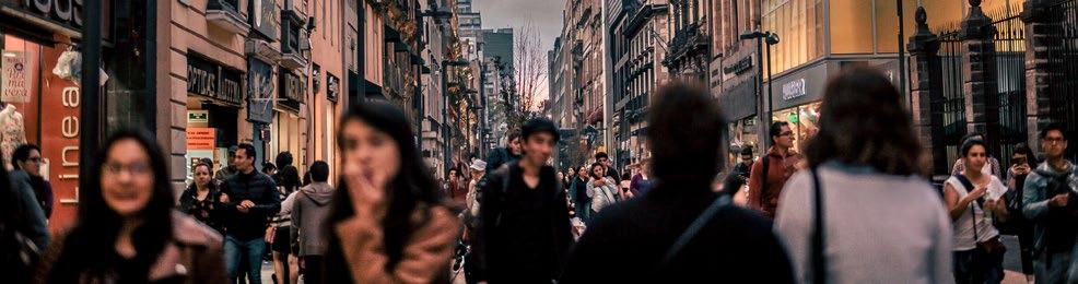 A panoramic view of a crowded city street with many pedestrians walking past shops and buildings under an overcast sky.