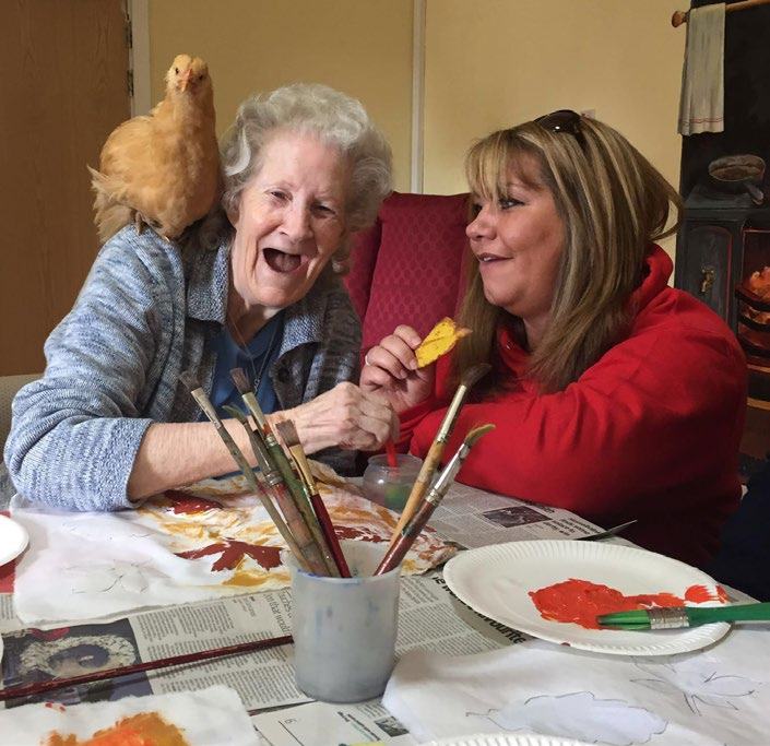 An older woman with a chicken on her shoulder and a younger woman smiling at a table with art supplies, engaged in a creative activity.
