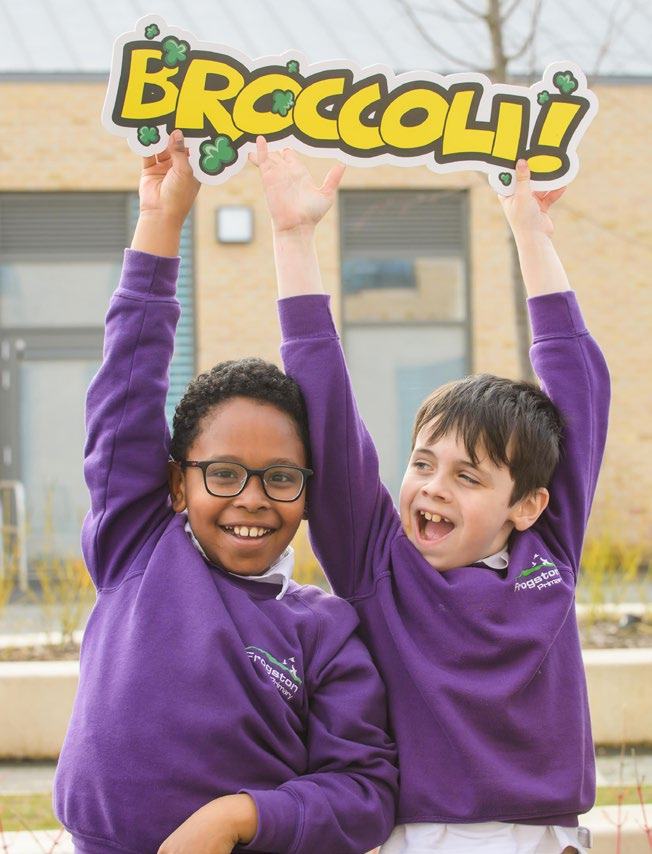 Two happy primary school boys in purple jumpers hold a sign overhead that says "BROCCOLI!" with decorative broccoli florets, likely promoting healthy eating.