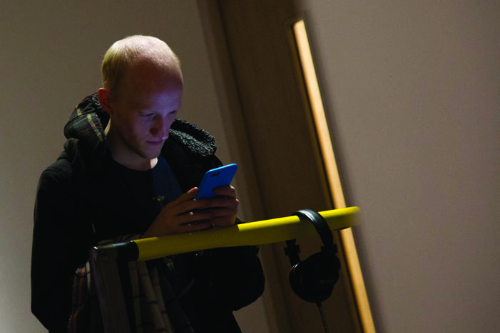 A person looking intently at a blue smartphone. Headphones hang from a yellow rail in the foreground, suggesting use in a gallery or public space.