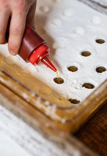A hand squeezes liquid from a red bottle into small indentations in a white powdered mold, part of a food preparation process.
