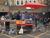 People gather around a food cart with a red umbrella in an urban outdoor setting, possibly a market or event.