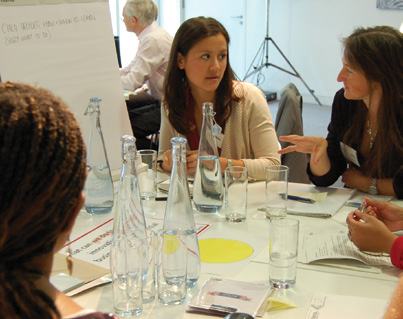 Participants in a workshop or meeting discussing ideas around a table with water bottles and papers, a whiteboard in background.
