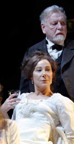 An actress in a white dress holds a glass, with an older male actor in period attire standing behind her.