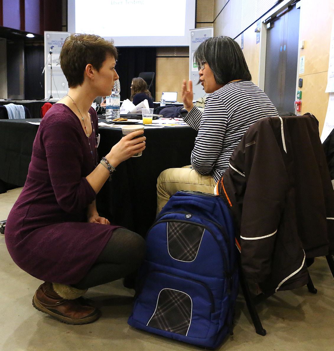 Two women in a conference setting are engaged in a discussion; one squats holding a coffee cup, the other sits and gestures.