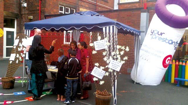 People gathered under a blue canopy tent at an outdoor event, with a large inflatable "your preston info" display nearby.