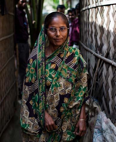 A woman in a patterned sari and glasses stands in a narrow alleyway between homes made of woven material.