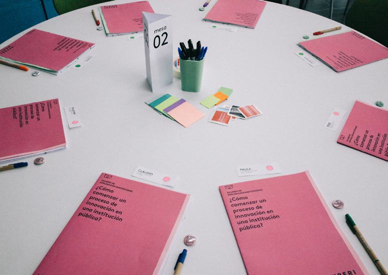 Overhead view of a workshop table set with pink folders, pens, and a "Mesa 02" sign, indicating a public institution innovation workshop.