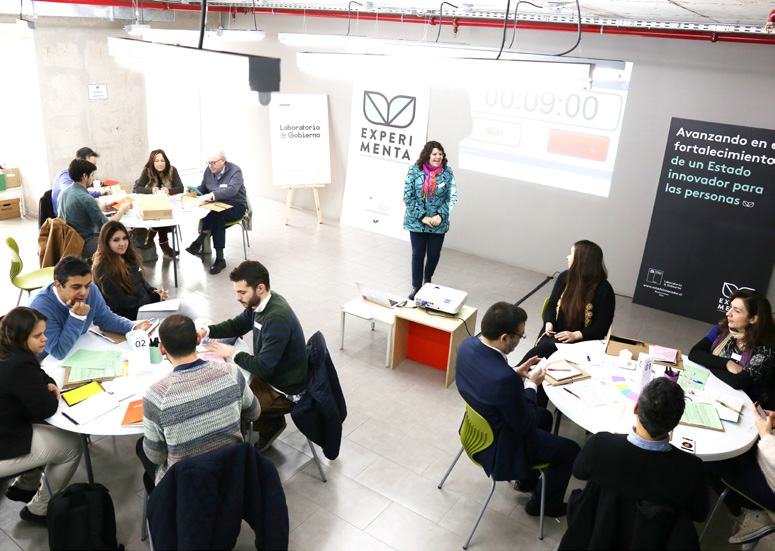 Participants at round tables during a workshop, with an instructor presenting and a screen showing a timer and "Laboratorio de Gobierno" branding.