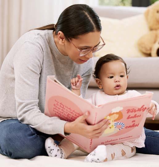 A woman in glasses and a baby sit on a rug, reading a pink children's book titled "Kirsty bakes a cake" together.