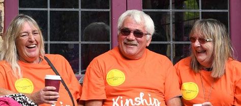 Three smiling adults wearing orange "Grandparents Plus" t-shirts, sitting together and laughing, possibly at an outdoor event.