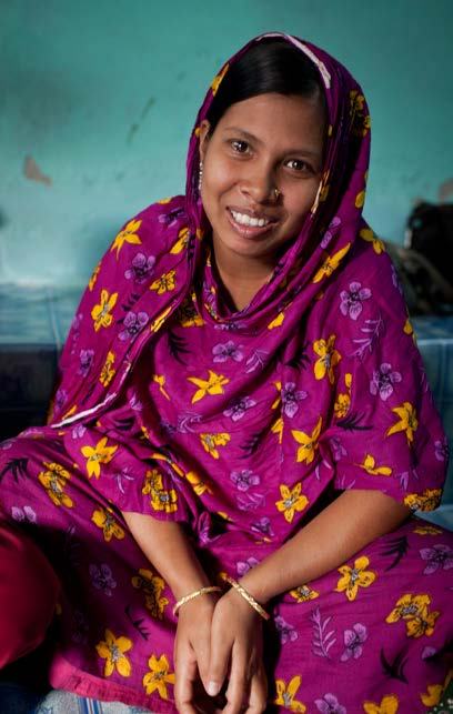 A smiling woman in a purple floral dress and bangles sits cross-legged on a mat, looking at the camera.
