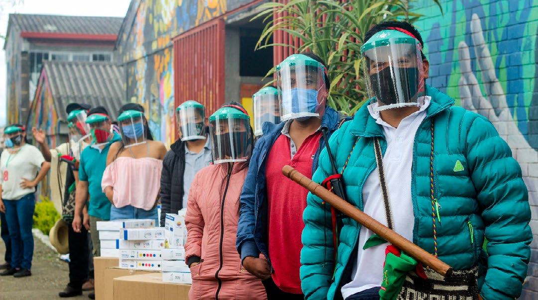 Group of people in face shields and masks lining up outdoors with boxes, possibly during a distribution event.