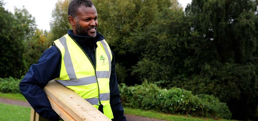 A smiling man in a yellow high-visibility vest with a tree logo holds a wooden plank in a park setting.