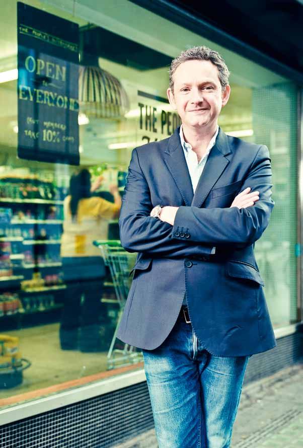 Man in a suit jacket and jeans smiles, standing outside "The People's Supermarket".