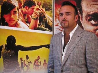 Man in a grey suit standing next to large movie posters, looking at the camera.