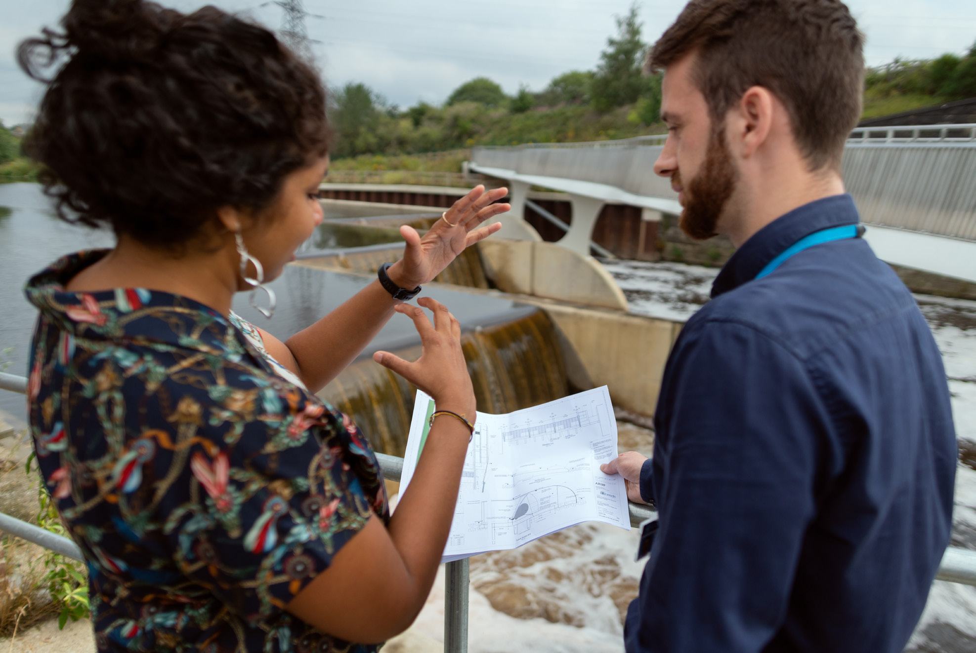 Two engineers or project managers discussing plans near a dam or water management structure, one holding blueprints.