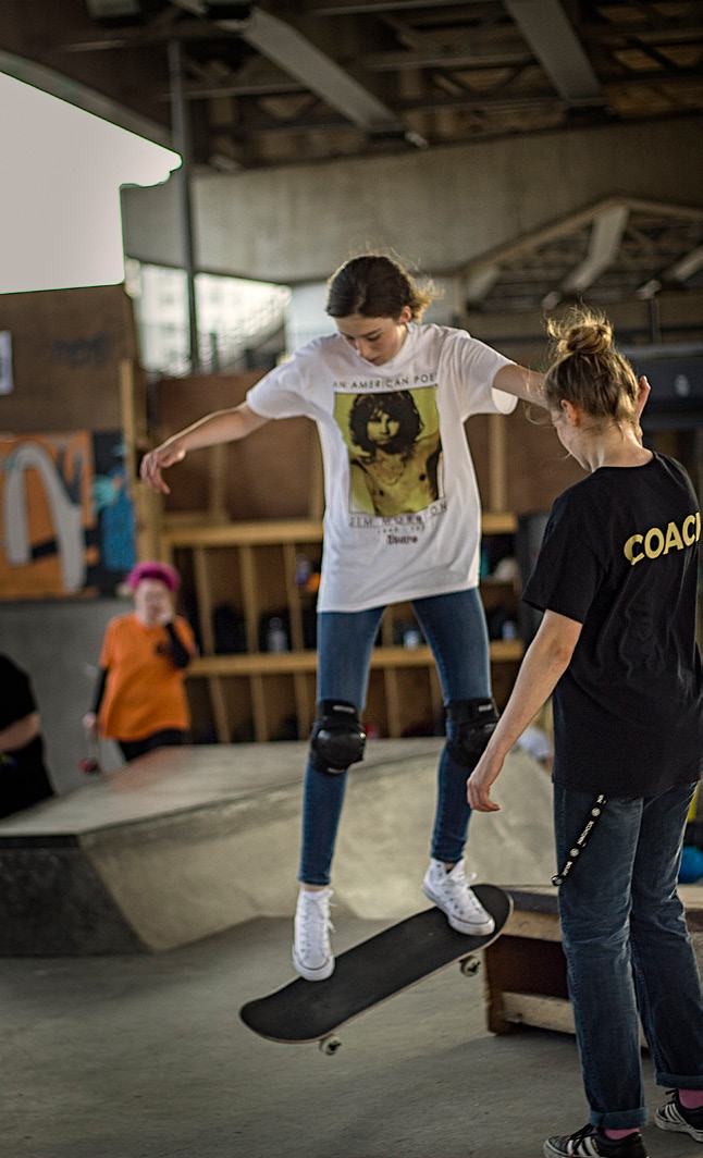 A young person in a white t-shirt and knee pads balances on a skateboard while an instructor in a black shirt stands behind them.