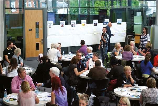 People conversing at round tables in a large room, with a historical timeline display on the back wall, suggesting a workshop or community meeting.